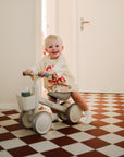 A smiling toddler rides the mushie Mini Toddler Bike on a red and white checkered floor.