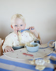 A young blonde-haired child sits in a high chair at a table set with a blue and white striped tablecloth and a mushie Silicone Placemat, using a fork to eat spaghetti from a blue bowl while wearing a bib for easy cleanup.