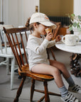 A young girl in a cream outfit and baseball cap sits on a wooden chair at a café table, eating an ice cream cone. Nearby are a children’s mug, eucalyptus vase, and the Bluey x Mushie Snack Cup by mushie—an essential for baby feeding.