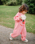 A young child in a pink strawberry-themed onesie walks outdoors on gravel, holding the mushie Bluey x Mushie Snack Cup and wearing white sneakers, with green grass and trees visible in the background.