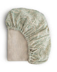 The mushie Extra Soft Muslin Crib Sheet, featuring a green and beige paisley pattern and an elastic edge, is shown partially folded on a neutral background—ideal for breathable baby bedding.
