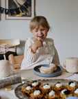 Smiling toddler enjoys a pastry, holding the mushie Snack Cup at the table.