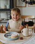 Toddler in mushie Silicone Baby Bib sits at table with food, donut, and a cup.