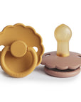 The FRIGG Daisy Natural Rubber Pacifier 2-Pack features two daisy-shaped pacifiers in golden yellow and light brown, each with scalloped edges, round handles, and natural rubber latex nipples. The FRIGG brand name is embossed on the front.
