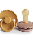 The FRIGG Daisy Natural Rubber Pacifier 2-Pack features two scalloped-edge pacifiers—one mustard yellow, one light brown. The left pacifier shows its handle; the right reveals its natural rubber latex nipple.