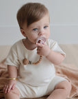 A baby with light brown hair and blue eyes, dressed in cream, sits on a beige blanket holding a FRIGG Moon Silicone Pacifier—one of the must-have baby essentials. A gold bracelet adorns their wrist as they look slightly to the side.