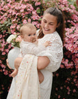 Woman holds baby with FRIGG Daisy Natural Rubber Pacifier near blooming pink flowers and a vase.