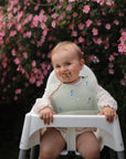 A baby sits in a white high chair, wearing a floral bib and holding the FRIGG Daisy Natural Rubber Pacifier from FRIGG, surrounded by vibrant pink garden flowers.