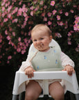 A baby sits outdoors in a high chair, wearing the mushie Bluey x Mushie Silicone Baby Bib. Pink flowers and green foliage bloom in the background, creating a cheerful, natural setting.