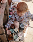A young child with light hair wears a mushie Long Sleeve Bib while playing with pastel toy cups and a teapot on a wooden surface outdoors.