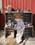 A young child in a mushie Long Sleeve Bib stands outside holding a cup beside wooden crates displaying toy dishes and potted plants, with a red wooden wall as the backdrop.