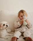 A young child sits on a cream-colored couch next to a small white dog, wearing a mushie Long Sleeve Bib and drinking from a light green bottle while looking forward.