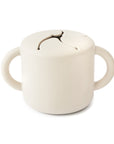 The mushie Snack Cup in Ivory is a cylindrical silicone cup with two side handles and a flexible, slotted lid for easy, spill-proof access—perfectly designed for young children.