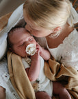 A young girl kisses a baby swaddled in tan with a FRIGG Daisy Natural Rubber Pacifier.