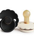 The FRIGG Daisy Natural Rubber Pacifier 2-Pack features two scalloped-edge pacifiers, one black and one white with a beige latex nipple, both standing upright on a white background.