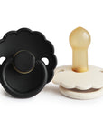 The FRIGG Daisy Natural Rubber Pacifier 2-Pack features two daisy-shaped pacifiers—one black, one white with a beige natural rubber latex nipple. The black pacifier faces forward; the white one rests on its side showing the nipple.