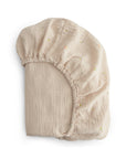 The mushie Extra Soft Muslin Crib Sheet is a beige fitted sheet with a subtle floral pattern and elastic edges, offering breathable comfort for baby bedding, shown on a white background.