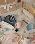 A flat lay of mushie's Stage 2 Blush Feeding Kit, featuring pastel silicone utensils, bowls, plates, a training cup with straw, spoons, and bibs, arranged with decorative ribbons and a gift box on a light background.