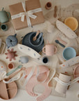 The mushie Sealife Bath Essentials Kit, featuring pastel-colored silicone bath items, is arranged on a white surface next to a beautifully wrapped gift box with a white ribbon.