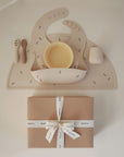 The mushie Stage 1 Bees Feeding Kit, in beige silicone, includes a bib, bowl, utensils, and cup on a printed mat. Below is a brown mushie gift box tied with a white ribbon.