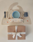 The mushie Stage 1 Dinos Feeding Kit—featuring a bib, spoon, fork, bowl, and cup with dino prints—is neatly arranged on a beige placemat above a gift-wrapped box tied with a white ribbon.