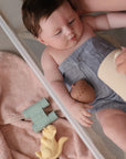 A baby in a small tub holds an acorn-shaped toy from mushie's Nature Mold Free Bath Play Set, covered with a towel, as an adult pours water from a jug. Nearby are other mold-free bath toys and toy binoculars from the set.