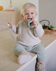 A smiling, blond-haired baby sits on a kitchen counter wearing a beige sweater and green pants, holding a FRIGG Lucky Symmetrical Silicone Pacifier 2-Pack (0-6 Months) that supports healthy oral development.