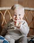 A smiling blonde baby sits on a wicker chair wearing a beige knit outfit, holding the mushie No-Drop Sensory Teether. Nearby, a blue and white blanket is draped.