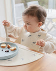 A young child in a mushie Silicone Baby Bib sits at a table indoors, holding a fork beside a blue plate with bread and small dark snacks. The child looks to the side and the scene is bright.