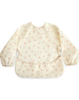 The mushie Long Sleeve Bib in Bows features a water-resistant cream design with elastic cuffs and a front pocket. This wipe-clean bib combines style and practicality, keeping your child tidy during mealtime.