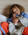 A young child with curly hair hugs a newborn on a bed, both in cozy pajamas. The mushie Silicone Pacifier Clip | Hera brings minimalist style to this warm moment as the baby rests peacefully, wrapped in a soft blanket.