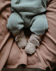A baby wears mushie Chunky Knit Pants in green organic cotton, paired with soft beige booties, while sitting on a light brown blanket. The baby's face is not visible.