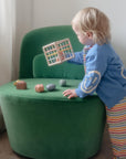Toddler plays with mushie Elastic Shape Sorter, sitting in a green chair in striped pants.