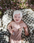 A smiling baby in a light brown outfit and white bonnet lies on a green and white checkered blanket outdoors, holding a FRIGG Daisy Natural Rubber Pacifier from the FRIGG Daisy Natural Rubber Pacifier 2-Pack. Pink petals and grass surround them.