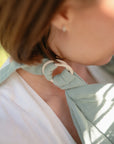 Close-up of a person wearing the mushie Muslin Nursing Cover ring sling in light green over a white shirt, with breathable fabric secured through a pale ring at the shoulder. The focus is on the ring and fabric; the face is partially visible.