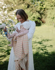 A woman in a white dress holds a baby wrapped in the mushie Organic Cotton Muslin Swaddle Blanket, standing near blooming tree branches on a sunny day.