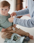 A young child sits on a white surface as an adult helps with the child's mint green shirt. Nearby is a light blue bag containing the FRIGG Lucky Symmetrical Silicone Pacifier 2-Pack (0-6 Months) from FRIGG for baby oral development.