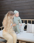 A woman with long blonde hair sits on a bamboo bench against a dark wood wall, holding a toddler in green knit clothes using the mushie Silicone Pacifier Clip | Luna. The bench is topped with patterned cushions.