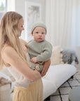 A woman with long blonde hair holds a baby in a light green knit outfit and hat, featuring the mushie Silicone Pacifier Clip | Luna, in a bright, cozy living room with white and beige decor.