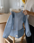 In a modern dining area with wooden furniture, someone holds up a mushie Long Sleeve Bib in light blue, highlighting its water-resistant design.