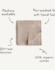The mushie Organic Cotton Muslin Swaddle Blanket, shown in beige with a folded corner, is 100% organic cotton, pre-washed for softness, machine washable, and designed for multiple uses as a swaddle or baby blanket.