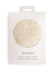 A white package with a circular window shows the beige Muslin Nursing Cover by mushie inside, labeled "mushie" and "Muslin Nursing Cover" in English, Spanish, and French to highlight its breathable design.