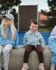 Three young children sit on a concrete bench outdoors. One child uses the FRIGG Daisy Night Silicone Pacifier from FRIGG, looking relaxed and happy with friends. A playground and trees can be seen in the background.