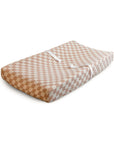 The mushie Extra Soft Muslin Changing Pad Cover, in a beige and white checkered pattern with two white safety straps, is displayed on a rectangular changing pad against a white background.