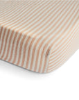 A close-up shows the mushie Extra Soft Muslin Crib Sheet, featuring beige and white vertical stripes, snugly fitted on a crib mattress against a white background.