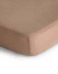 Close-up of a mattress corner covered with the mushie Extra Soft Muslin Crib Sheet in light brown, set against a white background, highlighting breathable baby bedding for comfort.