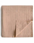 The mushie Organic Cotton Muslin Swaddle Blanket in Natural is neatly folded with a visible grid pattern, one corner turned up to show the underside, and displayed against a white backdrop.