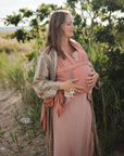 A woman in a peach dress stands among green plants, cradling her newborn in a matching peach carrier with a FRIGG Daisy Night Silicone Pacifier attached.