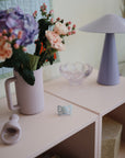 A pastel room features a ribbed vase of flowers, modern lavender lamp, glass dish, teether, and the FRIGG Butterfly Anatomical Silicone Pacifier 2-Pack (6-18 Months) by FRIGG on a light pink shelf—soft, cozy, and stylish decor elements.