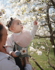 A woman holds a baby reaching for white blossoms, wearing a mushie Muslin Bib made from soft organic cotton, surrounded by bright flowers and greenery on a sunny day.